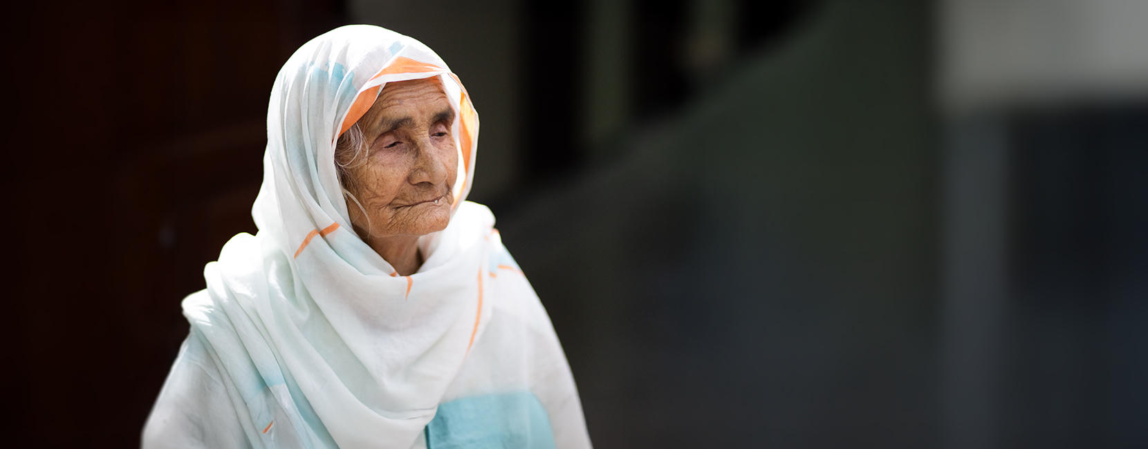 Elderly woman wearing a white headscarf with light blue and orange patterns, standing in natural light with a thoughtful expression.
