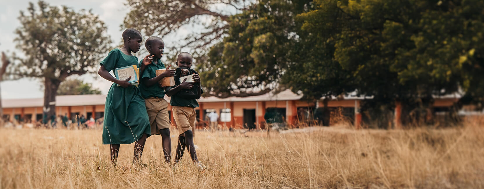 Three children in school uniforms walking together across dry grass, carrying books, with a school building and large trees in the background.