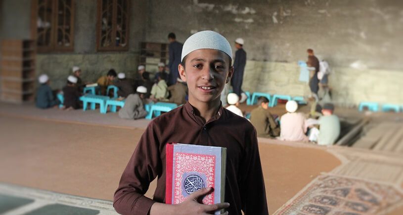 This image shows a young boy in traditional attire, smiling while holding a copy of the Qur'an. He is wearing a white cap and a brown outfit, and he is standing inside what appears to be a madrasa (Islamic school) or mosque.