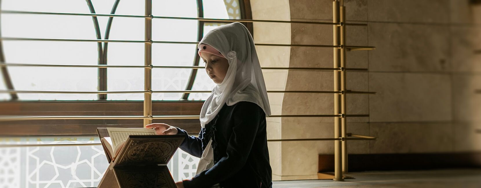 A young girl in a white headscarf and dark clothing reads the Quran in a well-lit mosque. She is seated by a large decorative window, focused on the open book placed on a wooden stand.