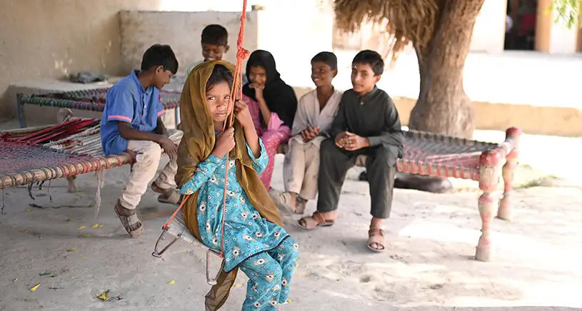 A young girl sits on a swing while other children sit nearby in a rural setting, supported through the OrphanKind programme by Penny Appeal.