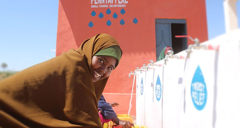 The image shows a woman in a brown hijab smiling while collecting water from a tap. She is at a water distribution point labeled with "Penny Appeal" and the slogan "Small Change, Big Difference."