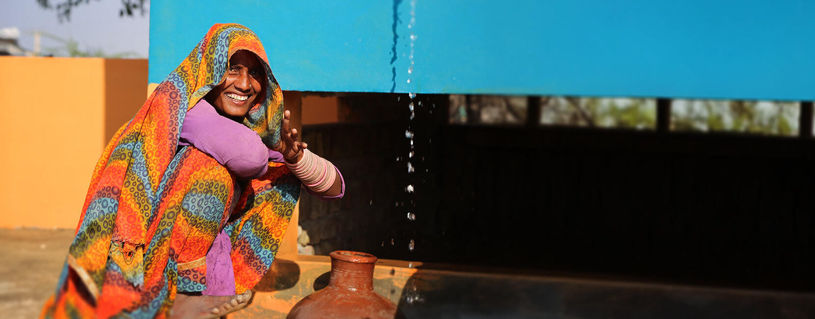 A smiling woman in a colorful sari collects clean water under a bright blue container, supported by Penny Appeal's Sadaqah Jariyah initiative to provide lasting benefits to communities.