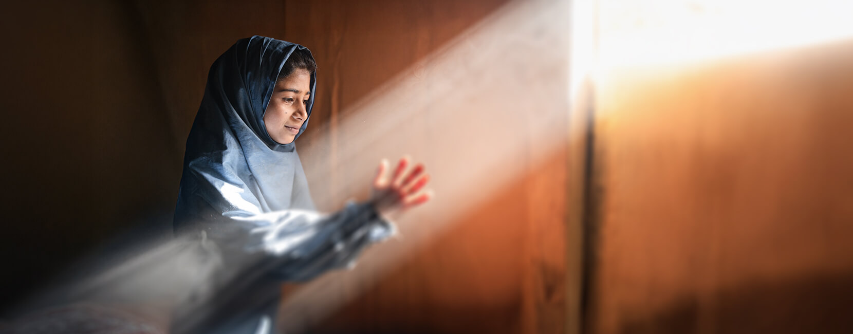 Young girl wearing a blue hijab holds her hands out towards a beam of light.