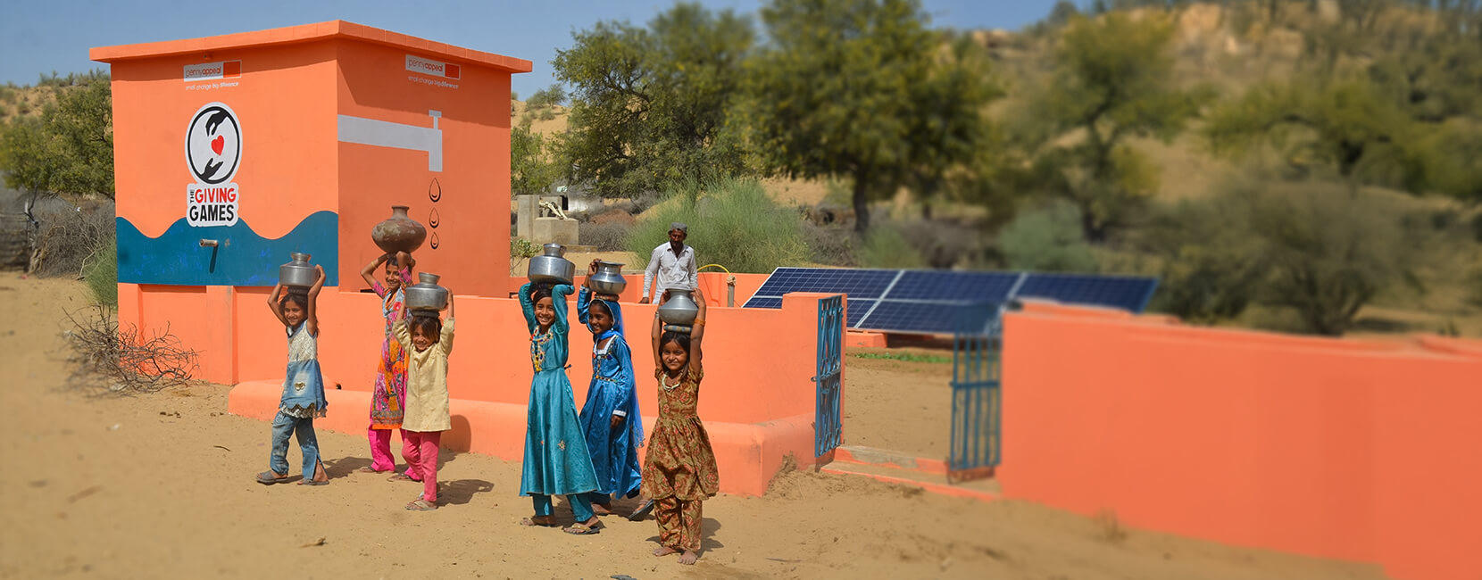 Group of smiling children carrying metal water containers on their heads, walking away from a bright orange community water facility in a desert area.