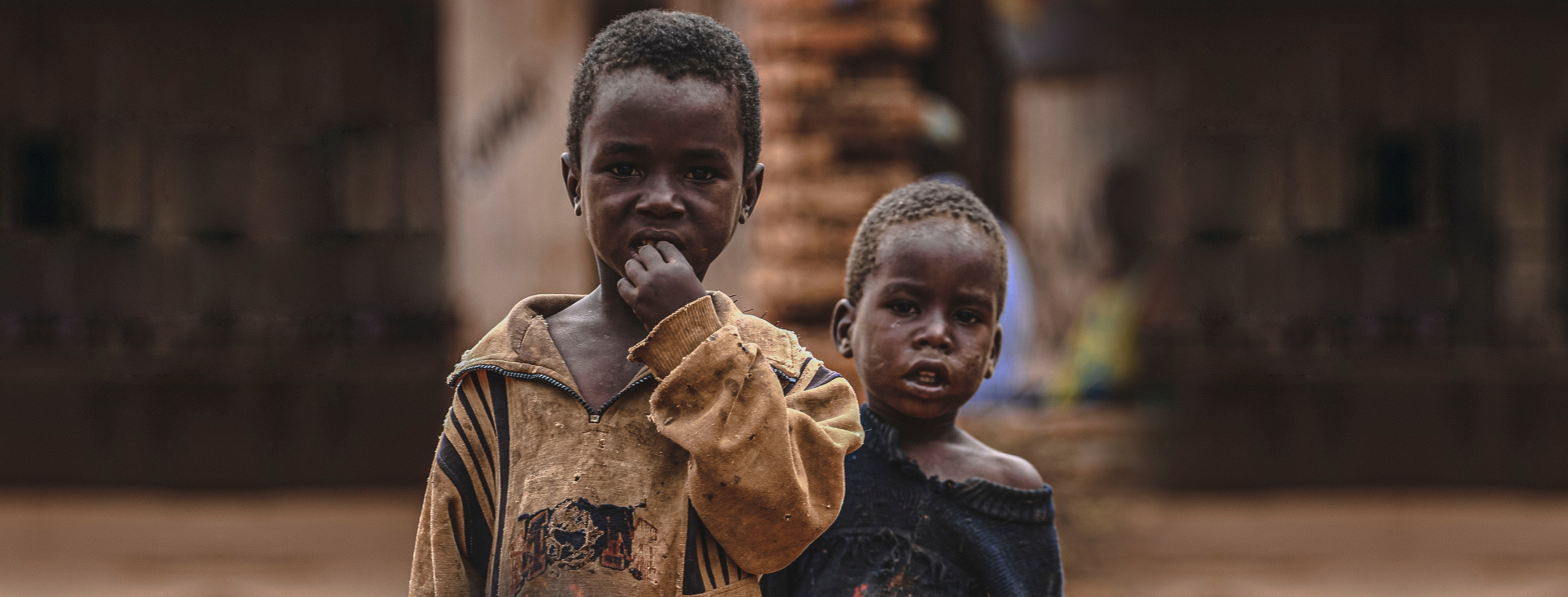 Two young children stand close together in a dusty street, wearing worn clothing, looking toward the camera. The image highlights the humanitarian crisis and urgent need for aid in the Penny Appeal Sudan Emergency campaign.
