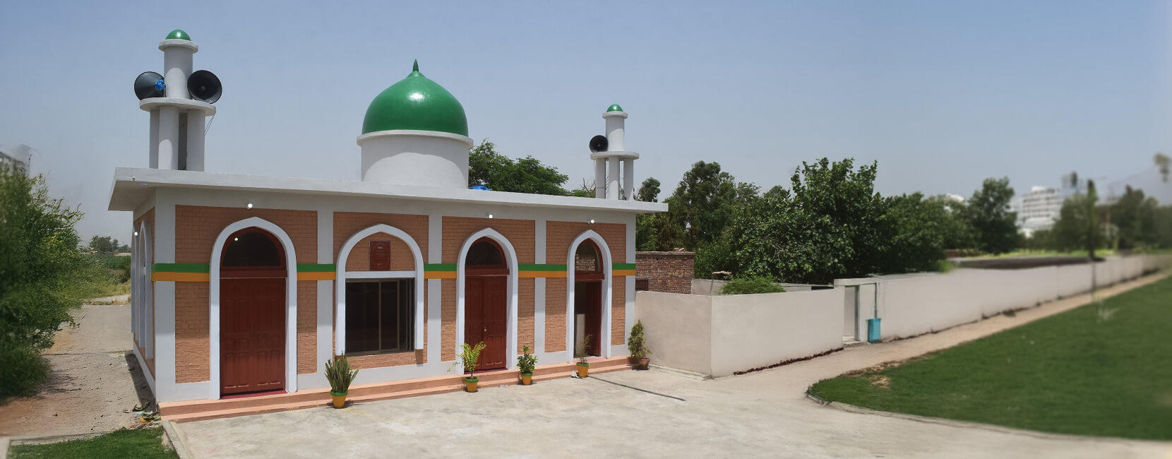 Newly constructed mosque with a green dome and twin minarets, surrounded by greenery on a sunny day.
