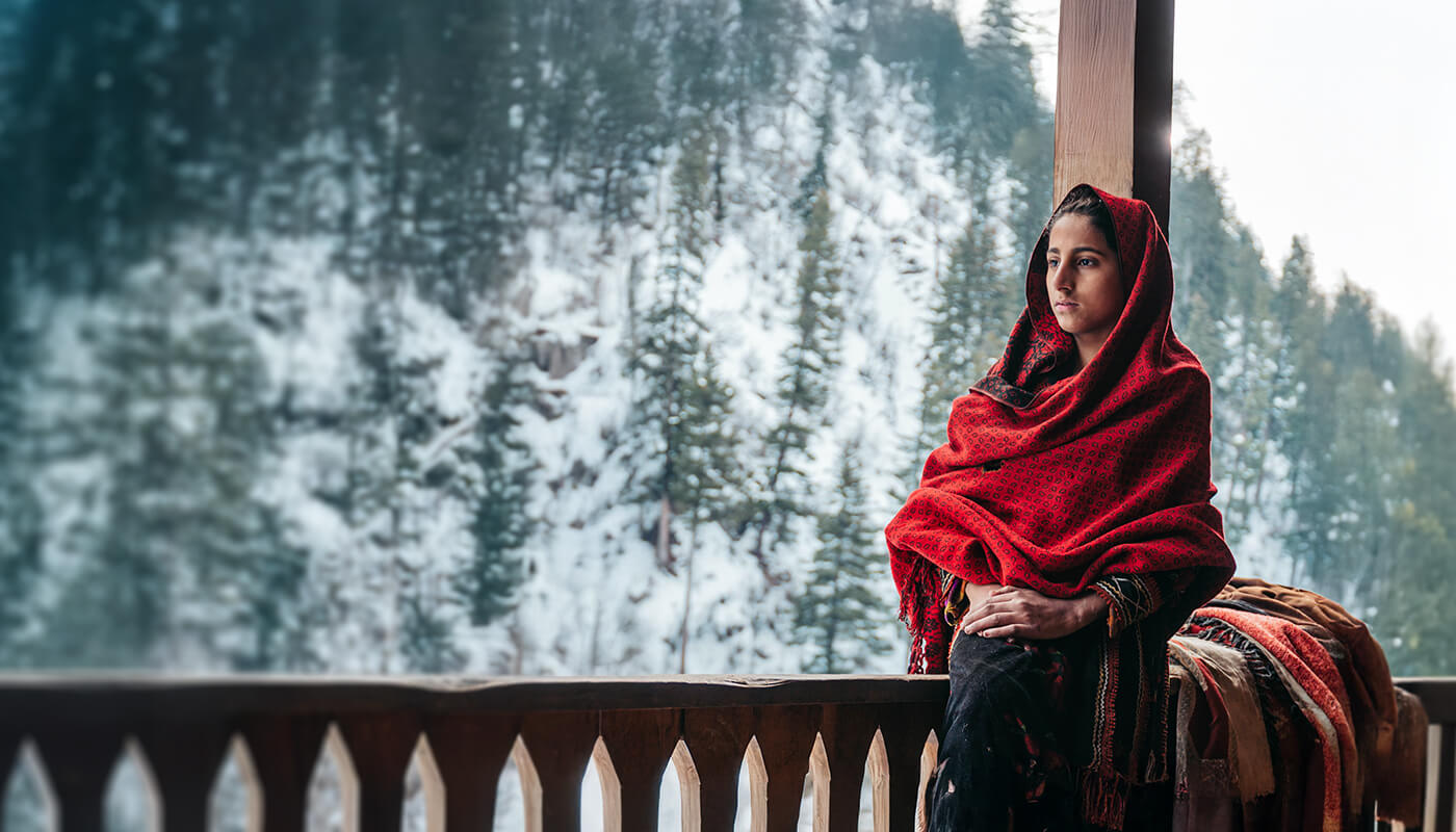 A person in a red shawl stands in front of a snowy forest backdrop.