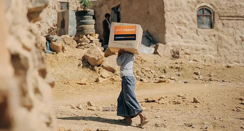 A person carries a Penny Appeal donated box of supplies through a sandy area.