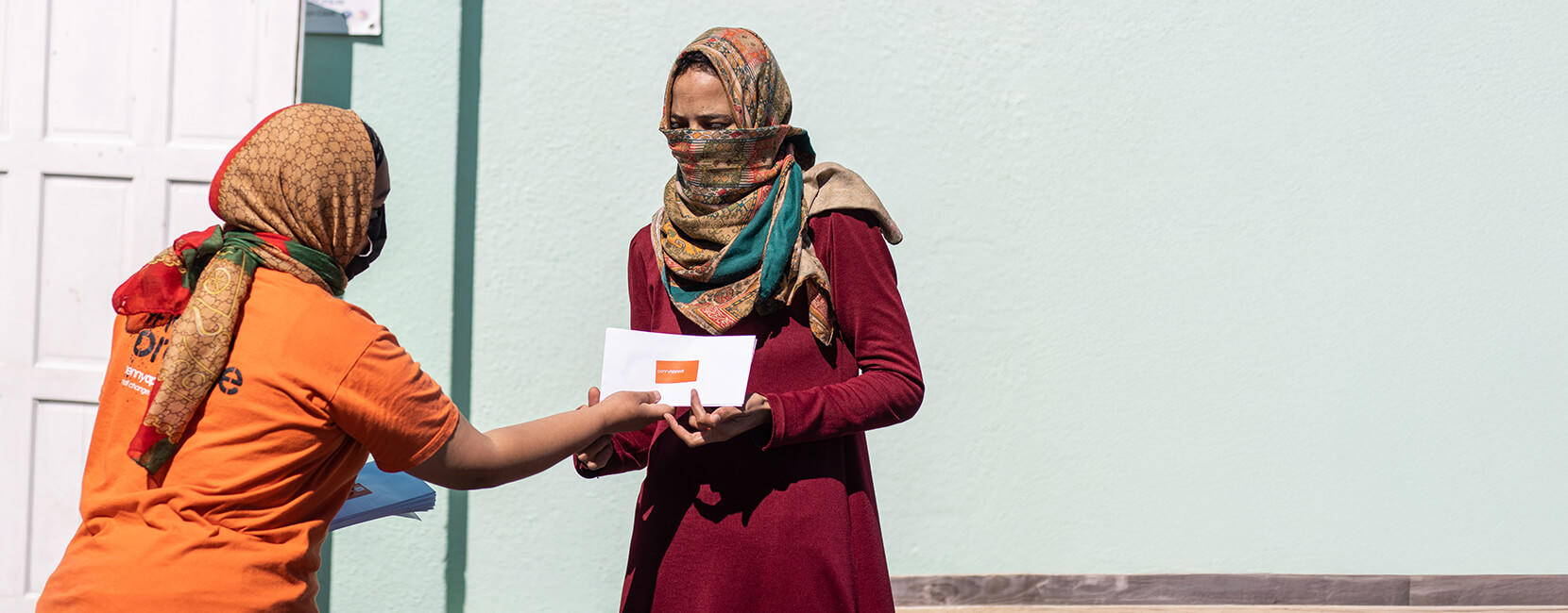 The image shows a woman in an orange Penny Appeal shirt handing an envelope to another woman wearing a maroon dress.
