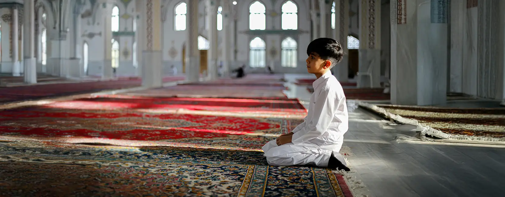 A young boy in white clothes kneels on a prayer mat.