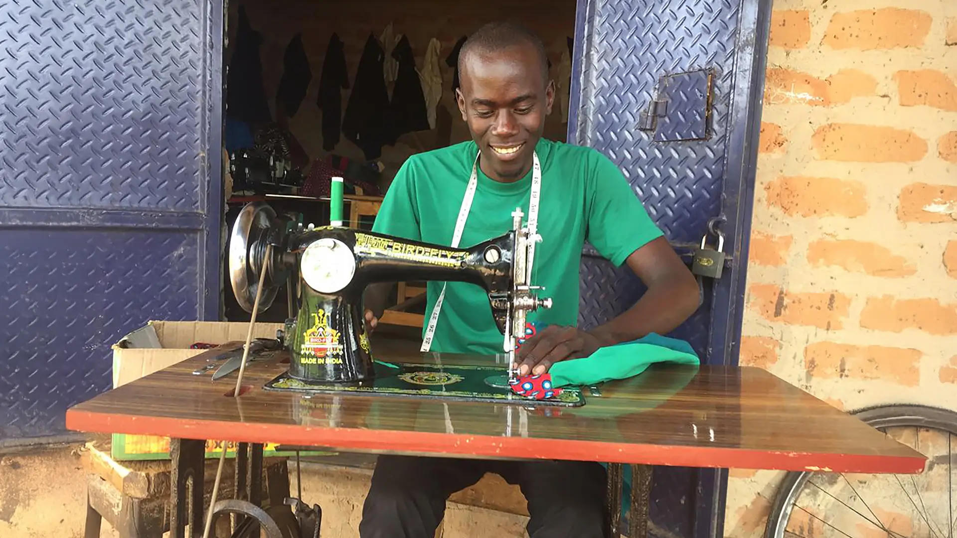 A smiling tailor works on a sewing machine at a wooden table, stitching colorful fabric in front of a workshop with blue metal doors and brick walls.