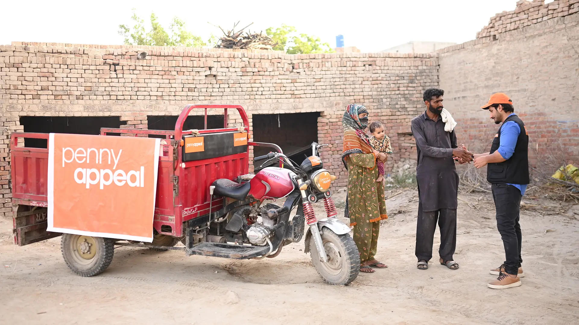 A Penny Appeal representative wearing an orange cap and vest shakes hands with a man standing beside a woman holding a child, next to a red motorbike with a Penny Appeal banner. The scene shows a community support initiative helping families in need.