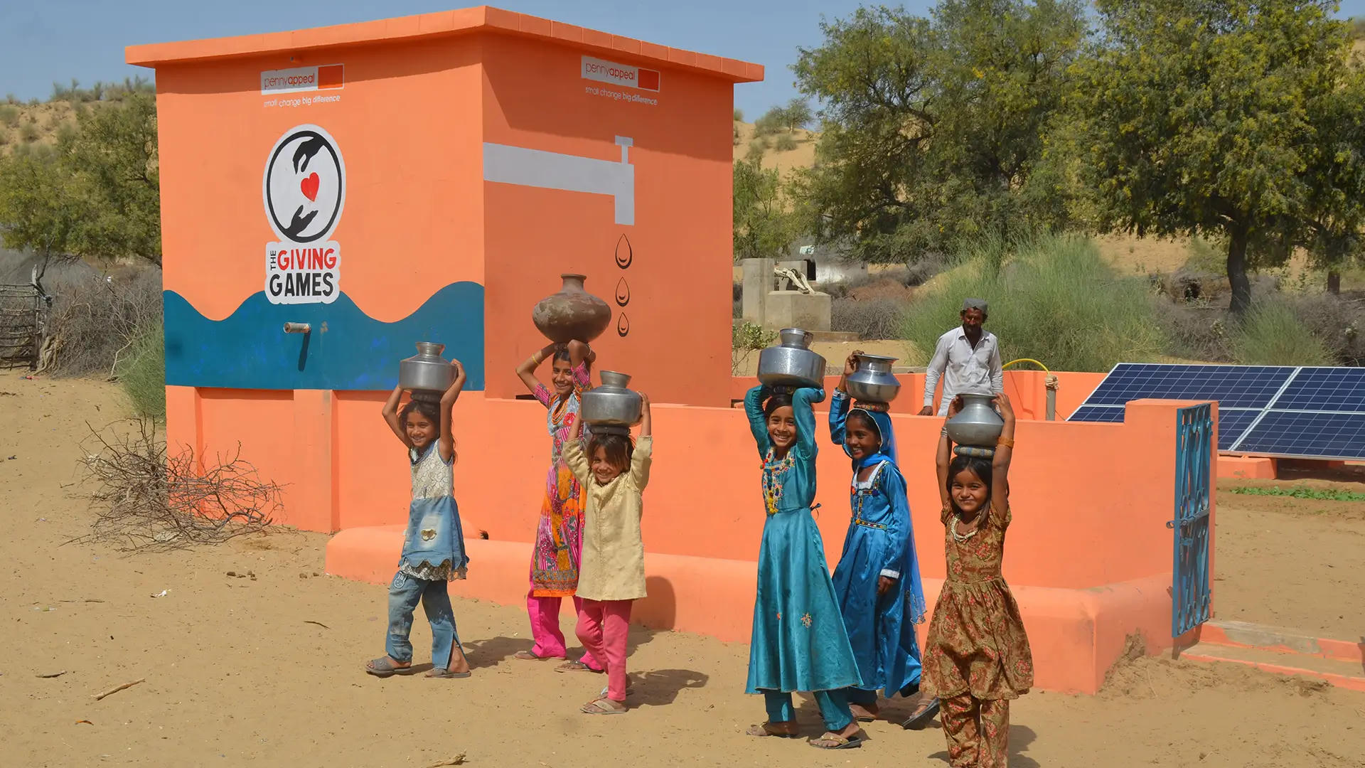 Smiling children carry metal pots filled with water from a Penny Appeal Thirst Relief Deep Well with solar panels and an orange water structure in the background.