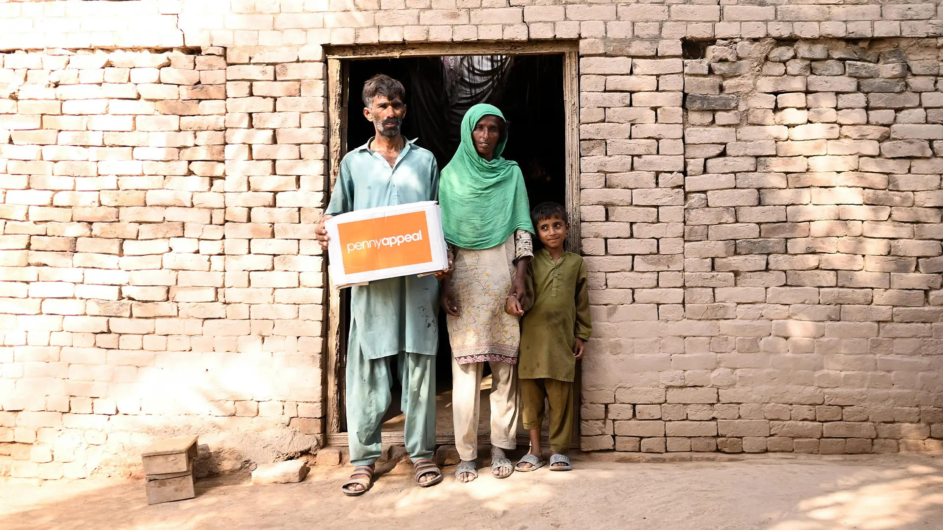 A family stands outside their home receiving a Penny Appeal Emergency Kit box, showcasing support for vulnerable communities in need.