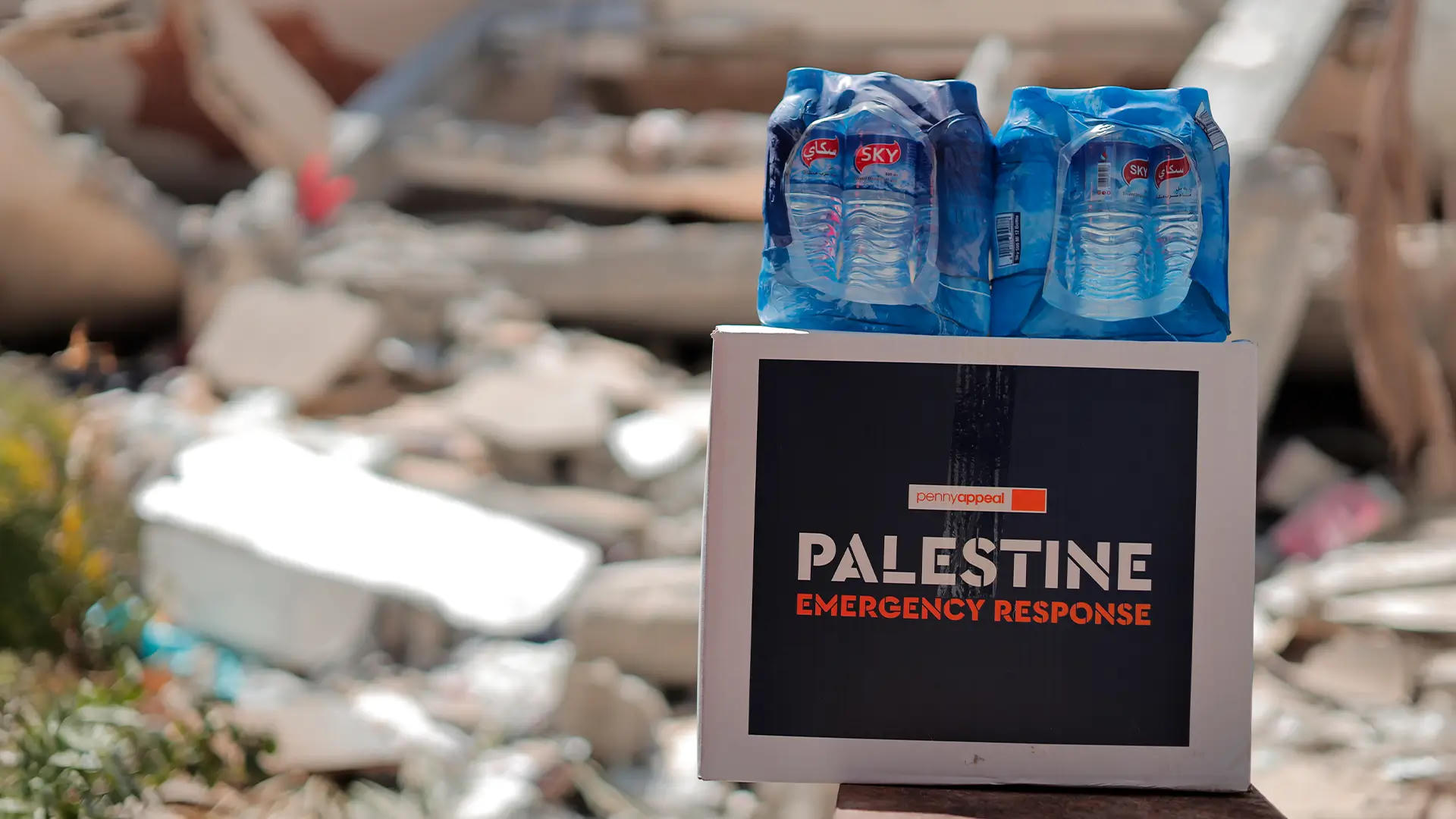 A Penny Appeal Emergency Kit labeled "Palestine Emergency Response" is topped with water bottles, placed amidst rubble, symbolizing urgent humanitarian aid in conflict zones.