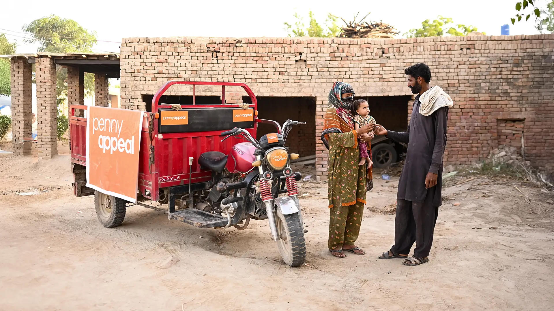 A woman holds an infant while stood talking to a man in front of a red rickshaw with a Penny Appeal logo on the side.
