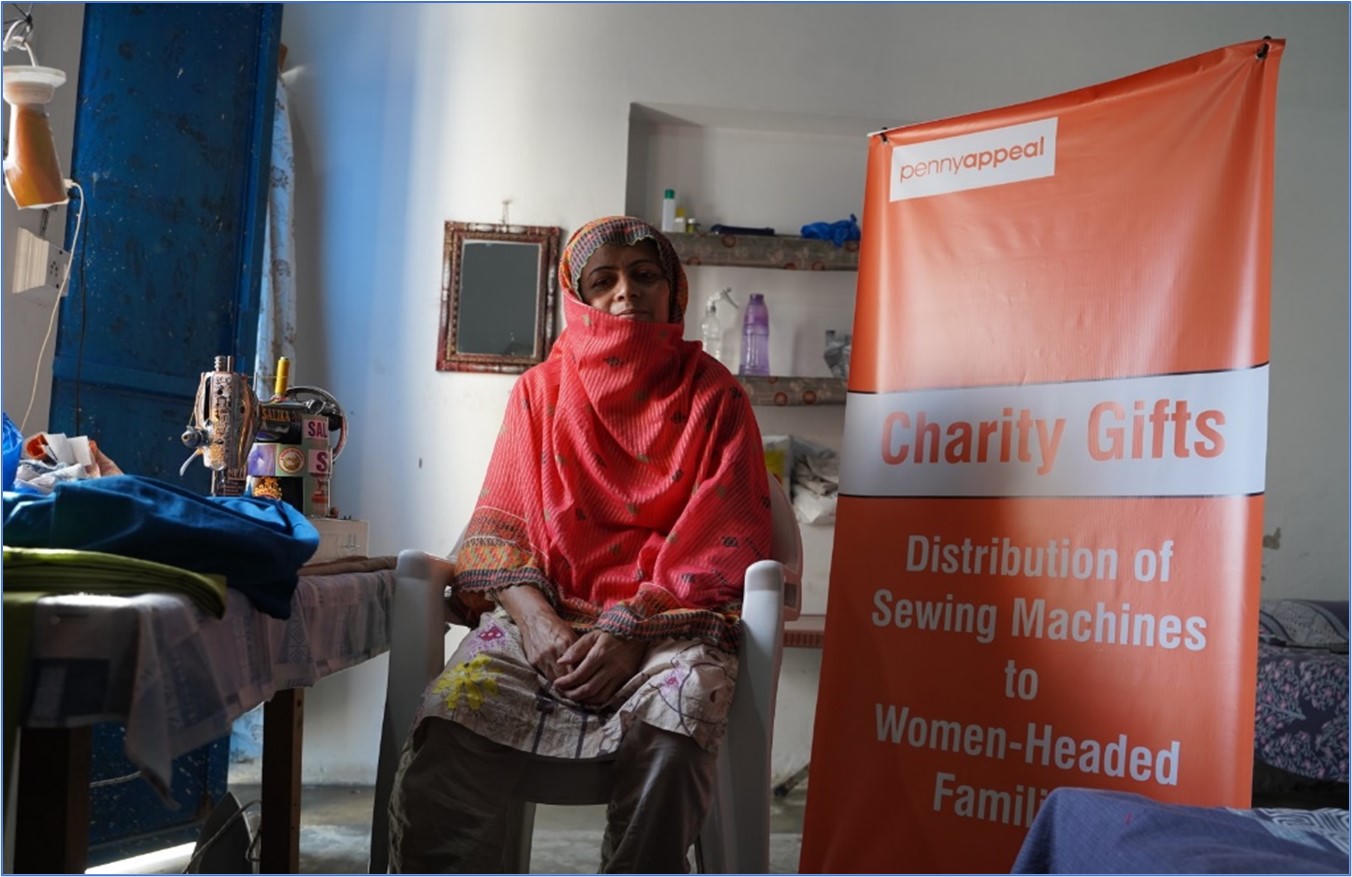 A woman sits in front of a Penny Appeal branded sign that says "Charity Gifts - Distributing Sewing Machines to Women-Headed Families"