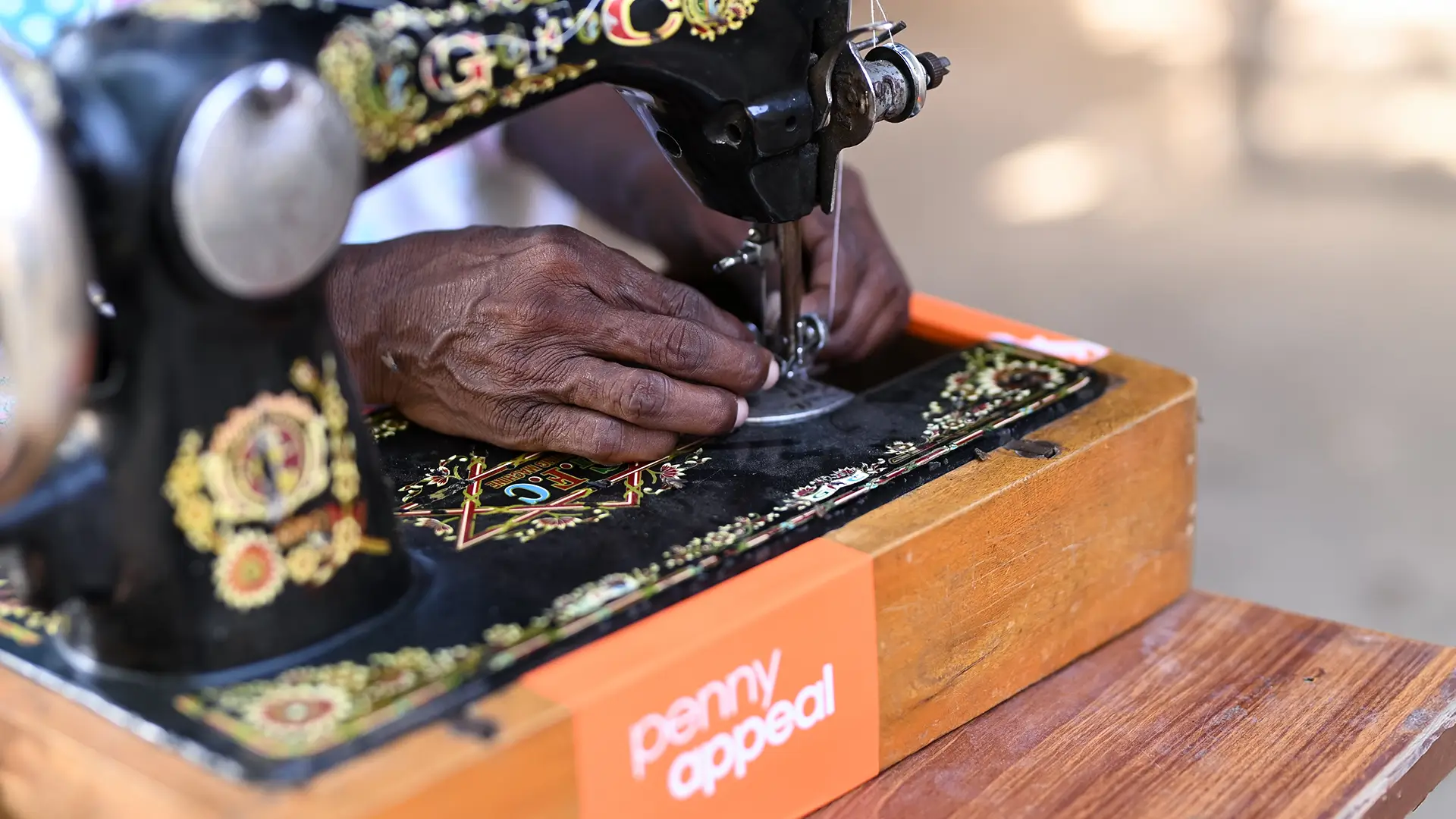 Hands use an ornate sewing machine with an orange Penny Appeal label on the front.