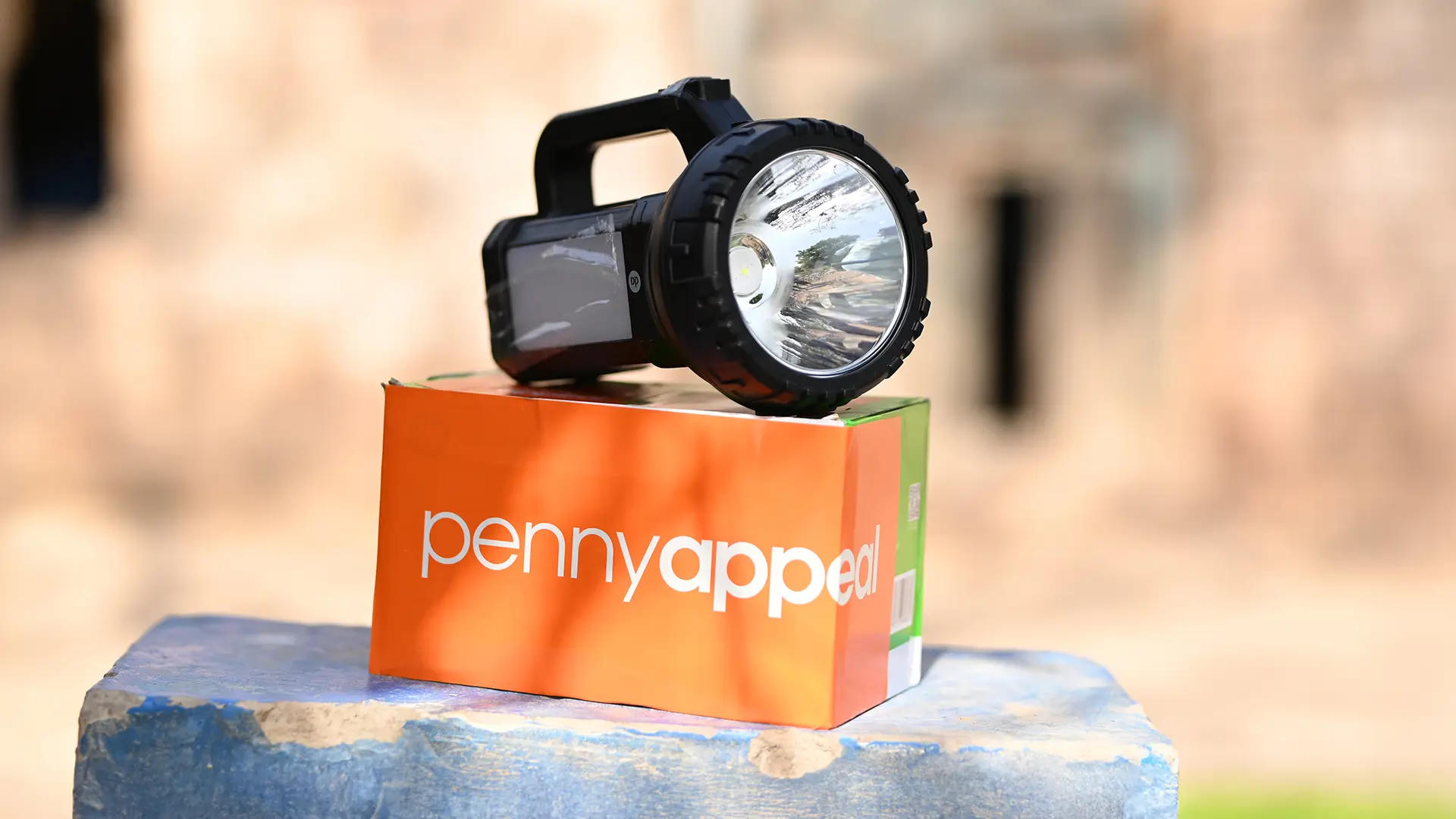 A solar lamp placed on a colorful box with the "Penny Appeal" logo visible. The lamp has a sturdy handle and a large, round light source, and the background is softly blurred.