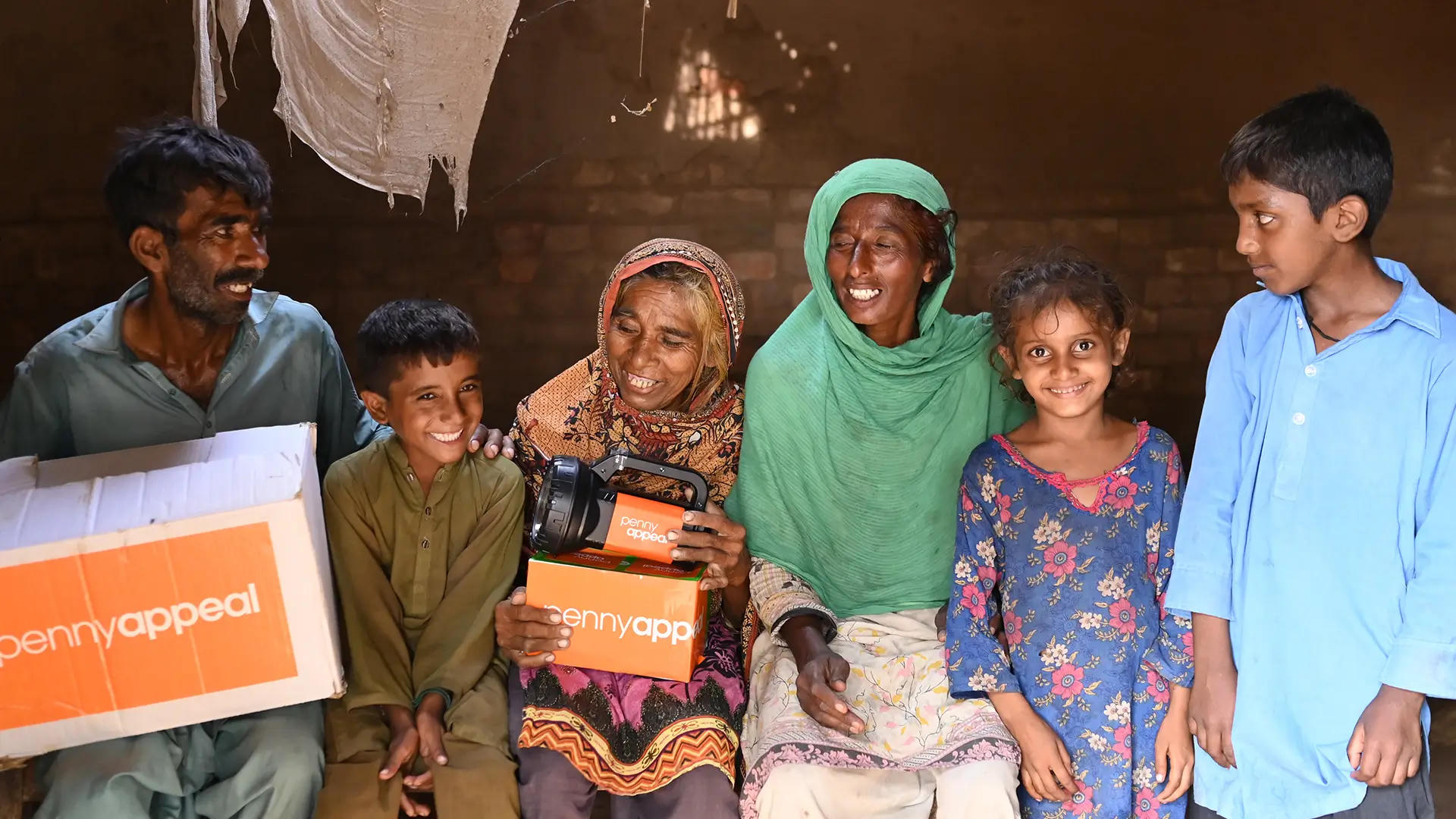 The image shows a group of six people, including children and adults, smiling and standing together in a dimly lit room. One woman holds a solar lamp with the "Penny Appeal" logo, while another person holds a box with the same logo..