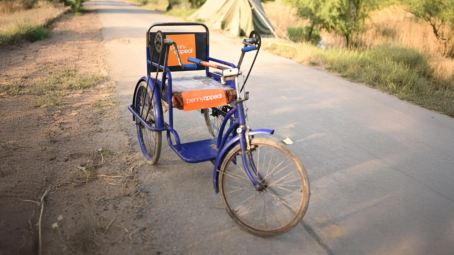 This image shows a wheelchair designed with a bike-like structure, also marked with the Penny Appeal logo. The wheelchair appears to be a specially designed model that can be pedaled, possibly making it easier for a caregiver or volunteer to move someone.