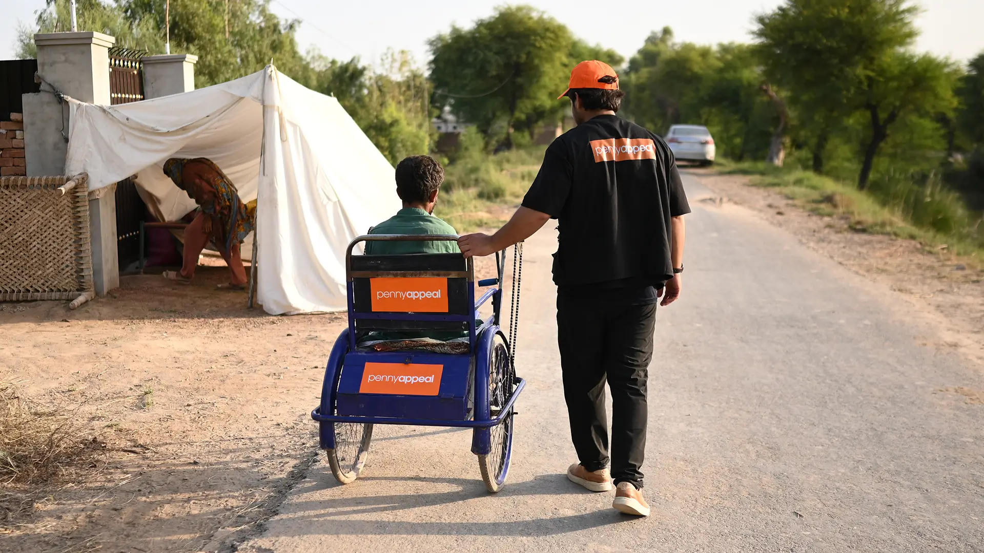 The image shows a volunteer from Penny Appeal assisting a person in a wheelchair along a rural road. The wheelchair is marked with the Penny Appeal logo, indicating the organization's involvement in providing aid.
