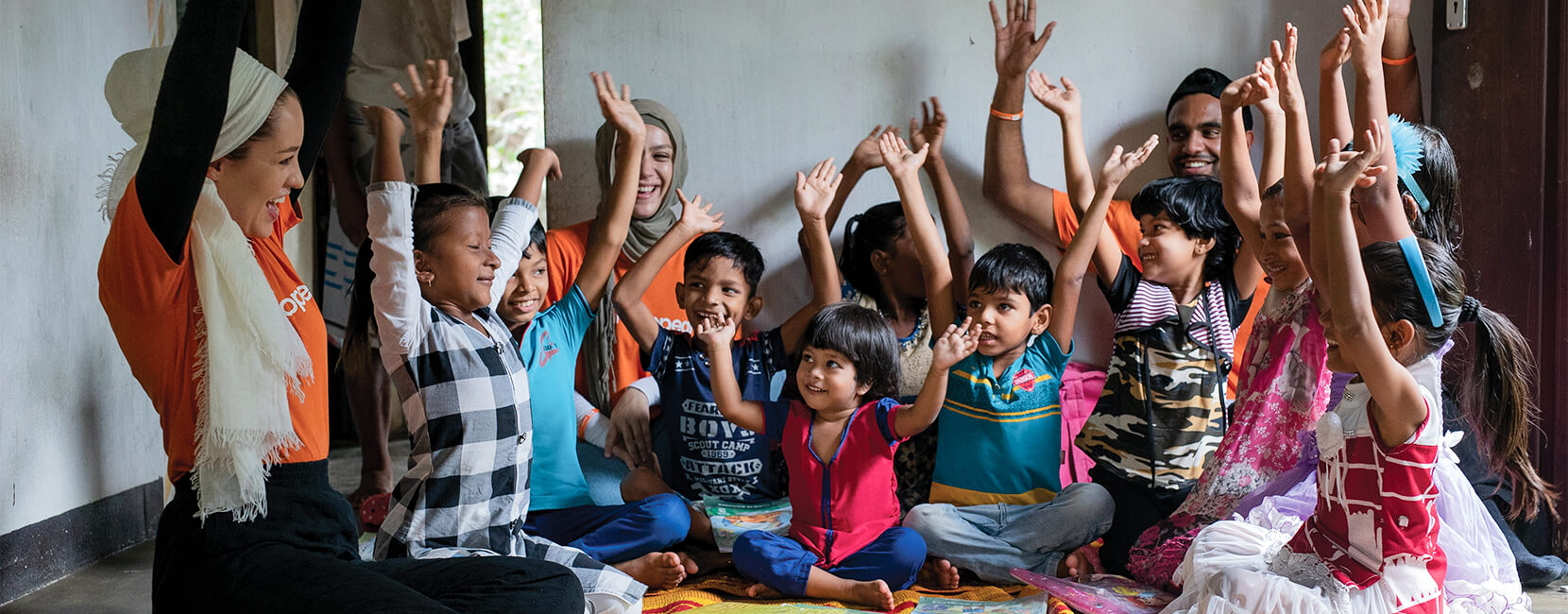 Group of children and volunteers sitting together indoors with raised hands during an educational or community activity.