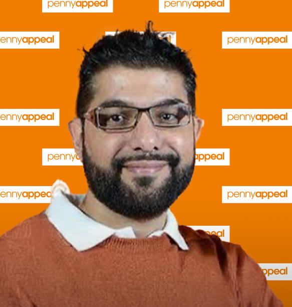 A close-up portrait of a man wearing glasses and an orange sweater. He is smiling in front of a backdrop with the Penny Appeal logo repeated.