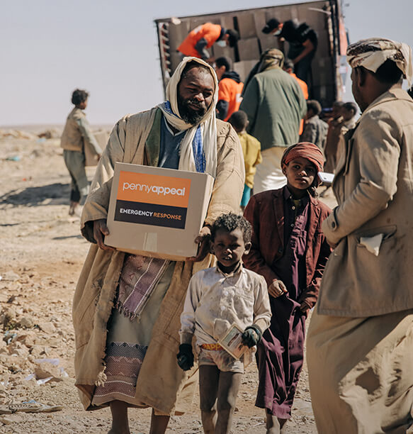 A woman with a tan hijab wearing an orange Penny Appeal shirt, seated and engaging in a conversation, with vintage decor and a clock visible in the background.