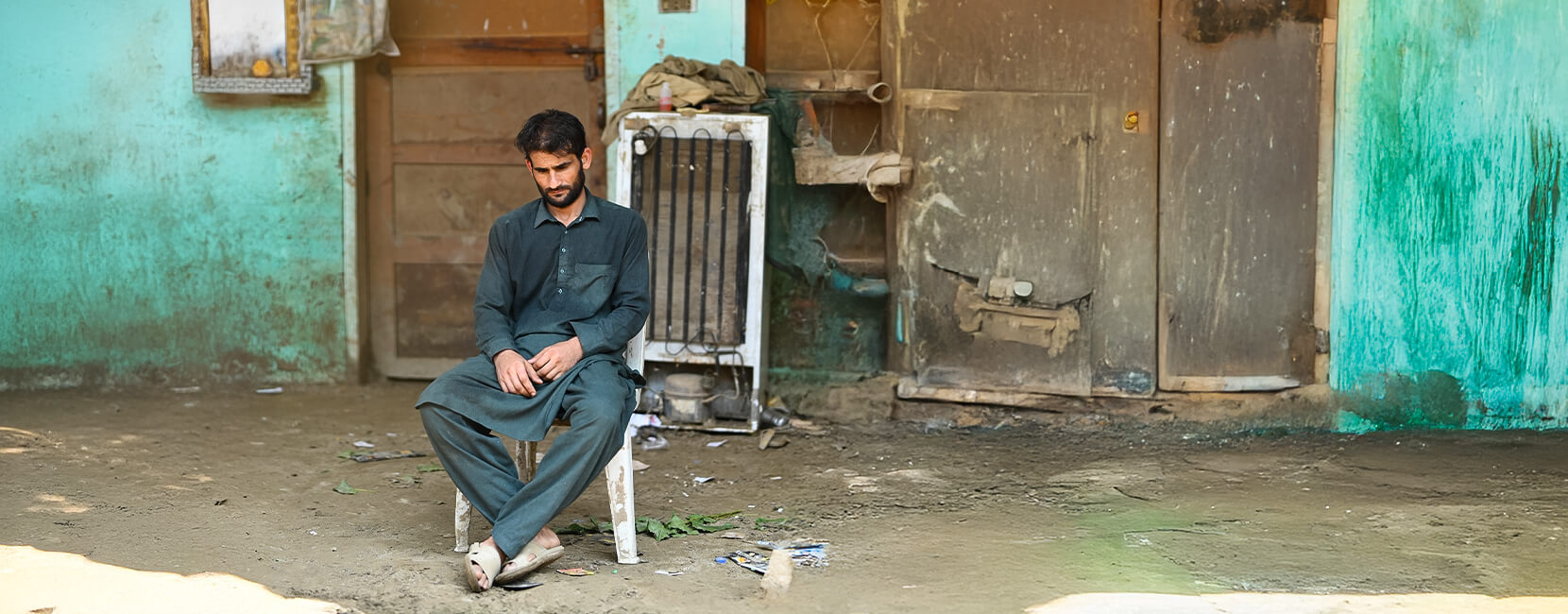 A man in dark traditional clothing sits alone on a plastic chair in front of a weathered building, looking down with a pensive or somber expression. The surrounding area appears dirty and run-down, with scattered debris and old appliances in the background.