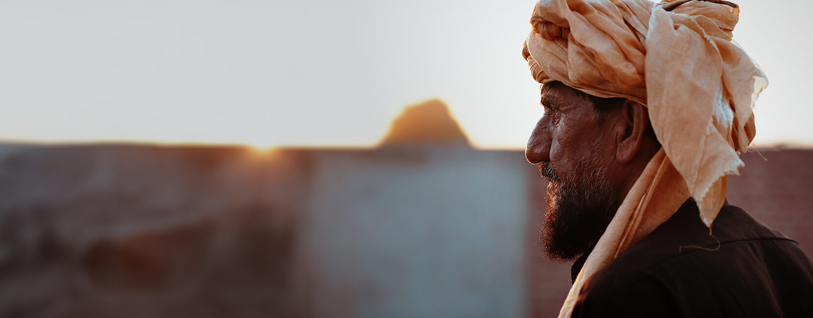Side profile of an elderly man wearing a traditional turban, standing outdoors at sunset with a calm expression, highlighting dignity and resilience.